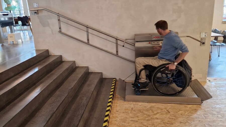 Un uomo è seduto su una sedia a rotelle. È in piedi davanti a una scala, sul sollevatore per sedie a rotelle e lo sta testando.