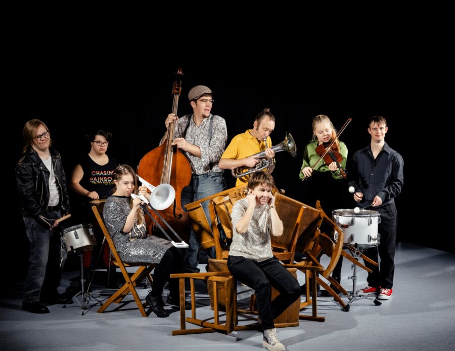 Otto attori del Komiktheater in azione con sedie e strumenti.