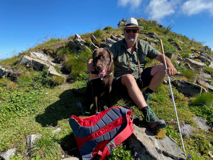 L'immagine mostra un uomo non vedente che fa una pausa con il suo cane guida durante un'escursione in montagna